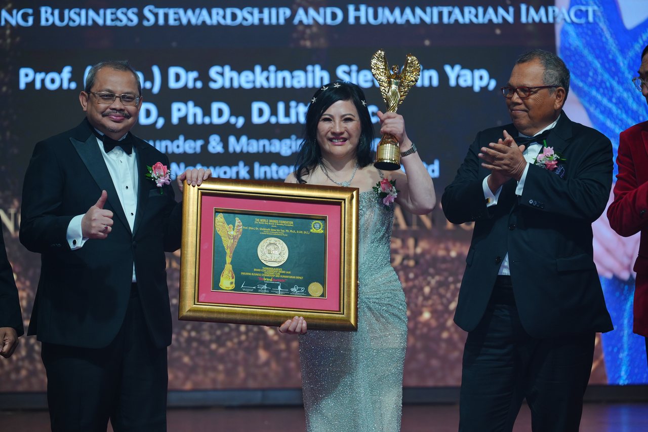 DR. SHEKINAIH SIEW SIN YAP, in a silver beaded gown, raises the gold winged trophy in her left hand and holds her framed certificate with a presenter giving a thumbs-up, as Tun Md Raus Sharif applauds, beneath the screen reading Enduring Business Stewardship and Humanitarian Impact