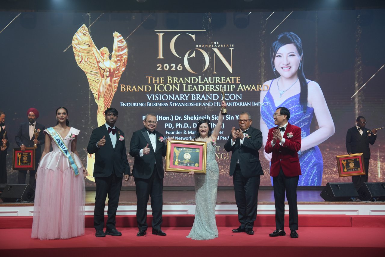 DR. SHEKINAIH SIEW SIN YAP raises the gold trophy at centre stage among the presenters, Prof. Dr. Edward Roy Krishnan and a presenter giving a thumbs-up at left, Tun Md Raus Sharif applauding beside her and Prof. Dr. KKJohan in a red velvet jacket at right, beneath the screen announcing Visionary Brand ICON, Enduring Business Stewardship and Humanitarian Impact, with her name and portrait