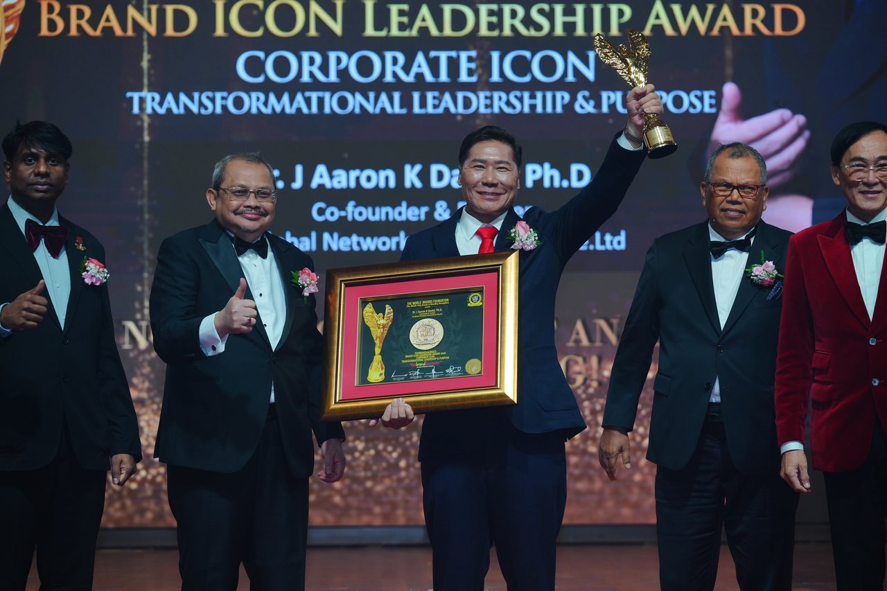 DR. J AARON K DAVID, in a navy suit and red tie, raises the gold winged trophy and holds his framed certificate as Prof. Dr. Edward Roy Krishnan and a presenter give a thumbs-up at left and Tun Md Raus Sharif stands beside him at right, beneath the screen reading Corporate Icon, Transformational Leadership and Purpose