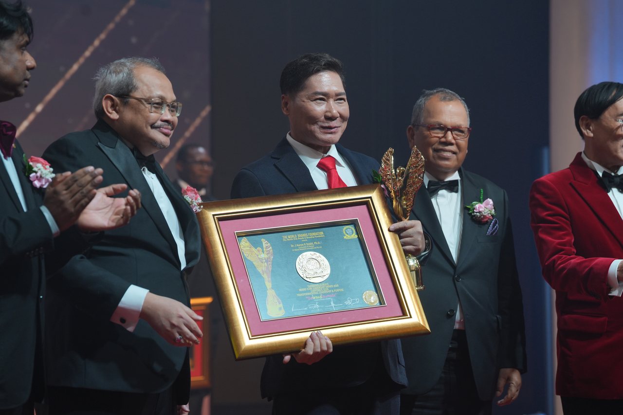 DR. J AARON K DAVID smiles holding his framed certificate in front of him and the gold trophy at his side, between presenters, as Prof. Dr. Edward Roy Krishnan applauds at far left and Tun Md Raus Sharif stands beside him at right