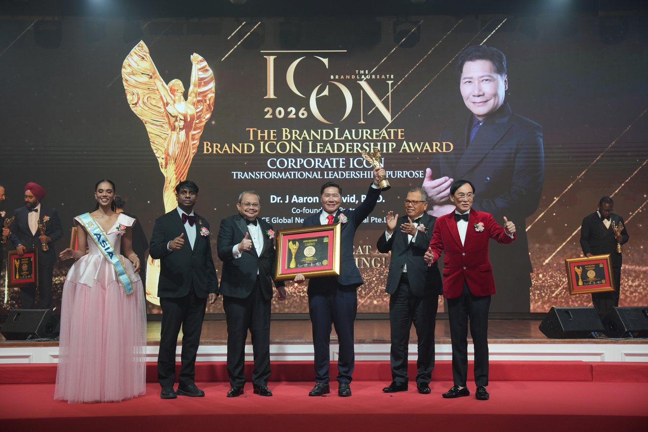 DR. J AARON K DAVID raises the gold trophy at centre stage beneath the screen announcing Corporate Icon, Transformational Leadership and Purpose, with his name and portrait, as Tun Md Raus Sharif applauds beside him and Prof. Dr. Edward Roy Krishnan gives a thumbs-up at left