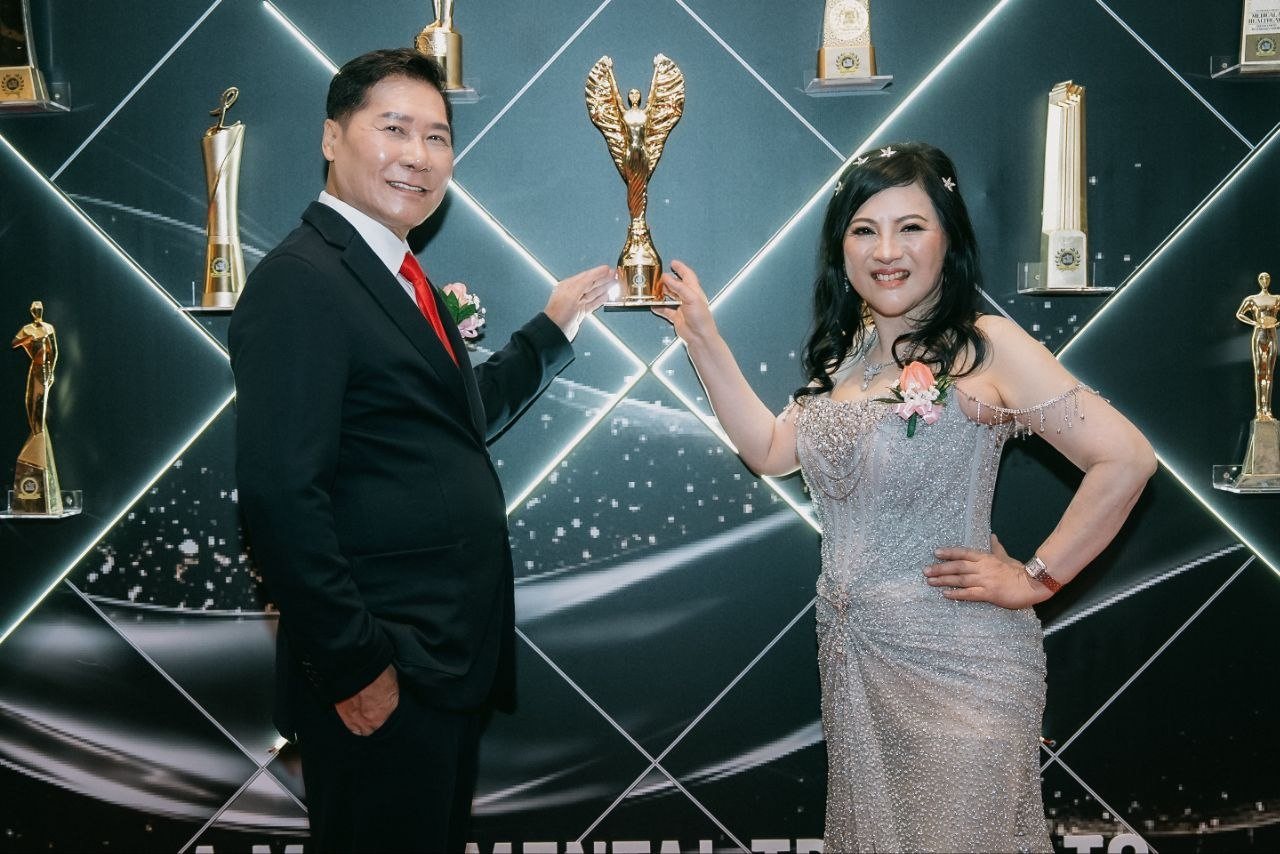 DR. SHEKINAIH SIEW SIN YAP, in a silver beaded gown, and DR. J AARON K DAVID, in a navy suit and red tie, hold one gold winged trophy up together in front of the illuminated awards wall, its black panels lined with trophies on glass shelves