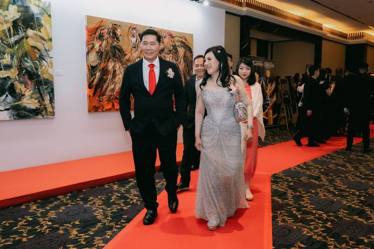 DR. SHEKINAIH SIEW SIN YAP, carrying a silver clutch, and DR. J AARON K DAVID walk the red carpet along a corridor hung with large paintings, one of horses, with guests following behind them