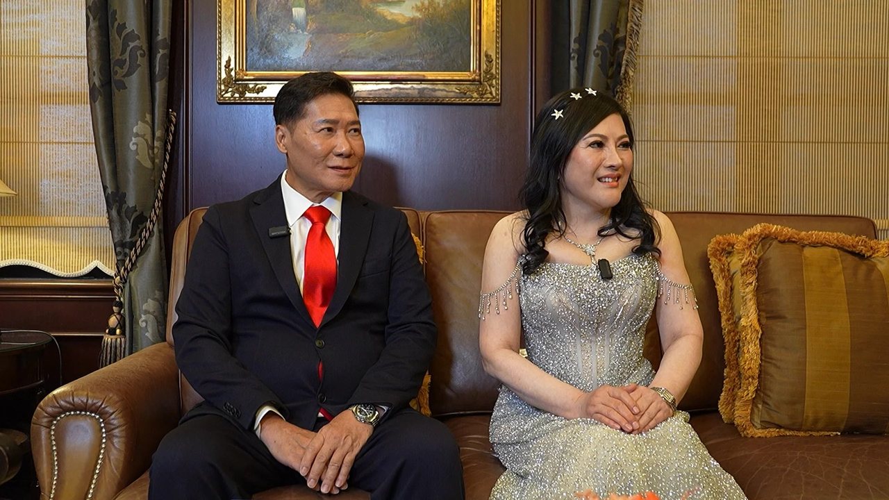 DR. J AARON K DAVID, in a dark suit and red tie, and DR. SHEKINAIH SIEW SIN YAP, in her silver beaded gown with star pins in her hair, wear lapel microphones as they sit side by side on a brown leather Chesterfield sofa beneath a gilt-framed landscape painting, looking towards an interviewer.