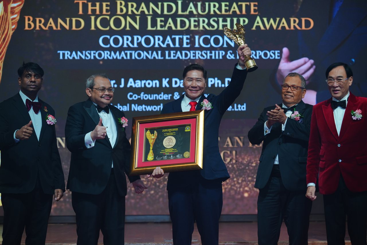 DR. J AARON K DAVID raises the gold trophy and holds his framed certificate as Tun Md Raus Sharif applauds beside him and Prof. Dr. Edward Roy Krishnan gives a thumbs-up at far left, beneath the screen reading Corporate Icon, Transformational Leadership and Purpose