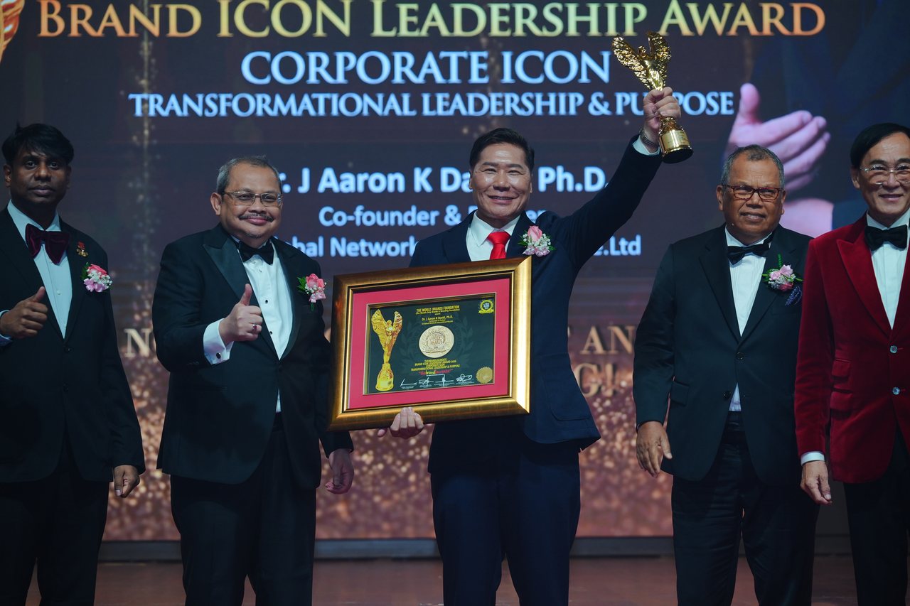 DR. J AARON K DAVID raises the gold winged trophy and holds his framed certificate as Prof. Dr. Edward Roy Krishnan and a presenter give a thumbs-up at left, with Tun Md Raus Sharif and Prof. Dr. KKJohan in a red velvet jacket at right, beneath the screen reading Brand ICON Leadership Award, Corporate Icon, Transformational Leadership and Purpose, with his name