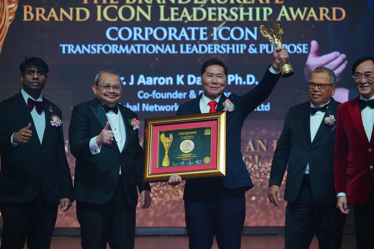 DR. J AARON K DAVID raises the gold winged trophy with his framed certificate held in front of him as Prof. Dr. Edward Roy Krishnan and a presenter give a thumbs-up at left and Tun Md Raus Sharif stands beside him, beneath the screen reading Corporate Icon, Transformational Leadership and Purpose