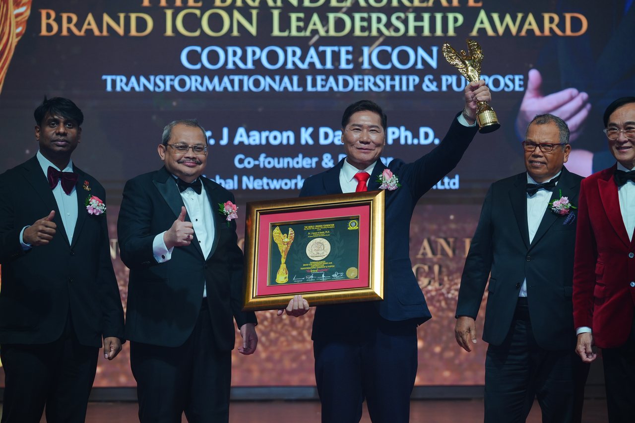 DR. J AARON K DAVID raises the gold trophy and holds his framed certificate among the presenters, Prof. Dr. Edward Roy Krishnan and a presenter giving a thumbs-up at left and Tun Md Raus Sharif and Prof. Dr. KKJohan at right, beneath the screen reading Corporate Icon