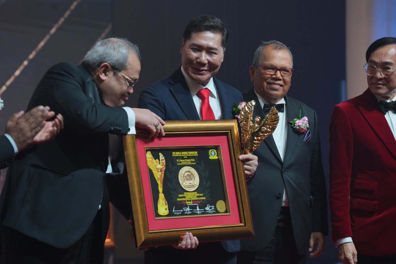 A presenter hands the framed certificate to DR. J AARON K DAVID, who holds the gold trophy, as Tun Md Raus Sharif smiles beside him and Prof. Dr. KKJohan looks on at right