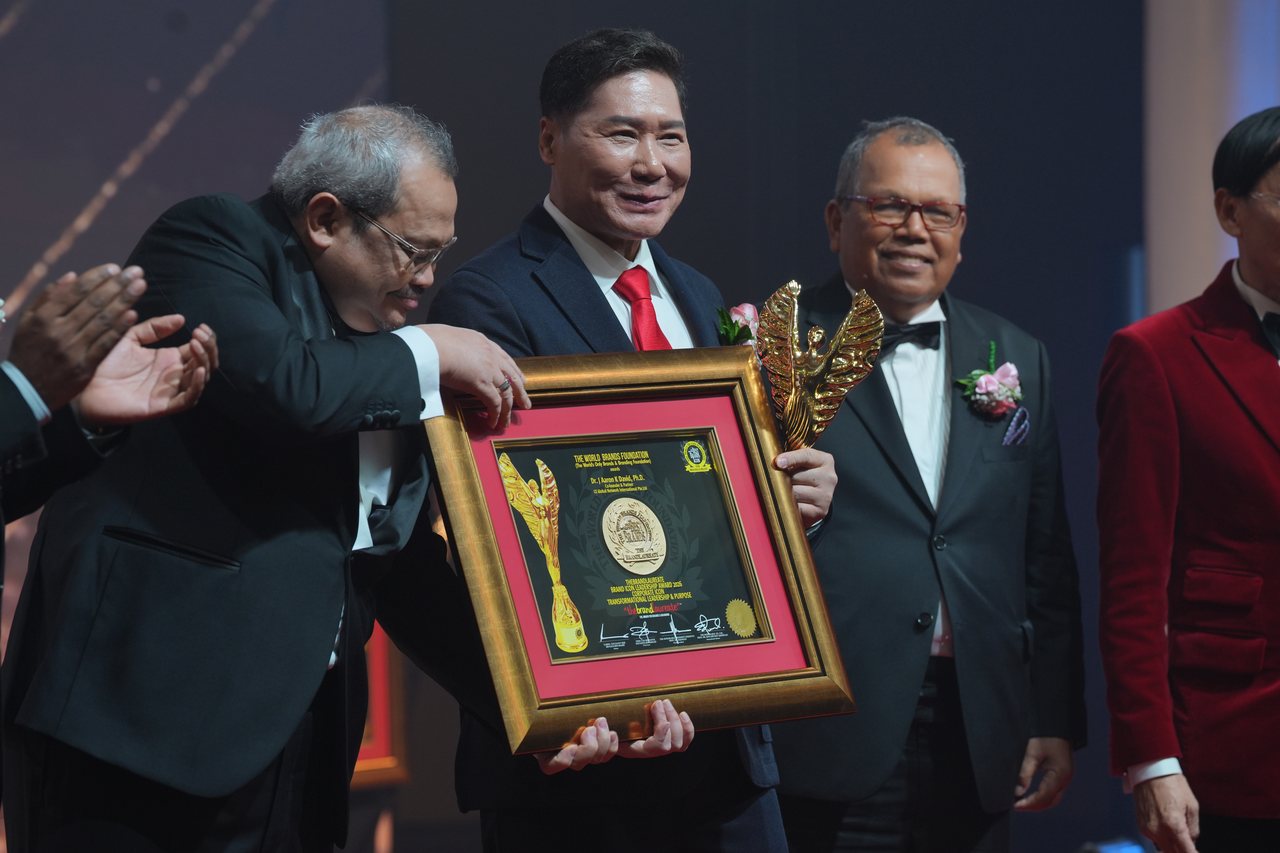 DR. J AARON K DAVID smiles as a presenter hands him his framed certificate, the gold trophy at his side, while Tun Md Raus Sharif smiles beside him at right