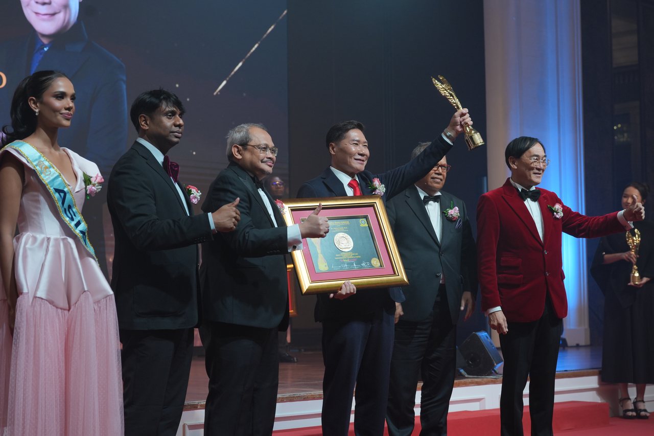 Left to right, Miss Universe Malaysia in a pale pink gown and sash, Prof. Dr. Edward Roy Krishnan and a presenter giving a thumbs-up, DR. J AARON K DAVID raising the gold winged trophy above his framed certificate, Tun Md Raus Sharif standing beside him, and Prof. Dr. KKJohan in a red velvet dinner jacket giving a thumbs-up, with a guest in black holding a trophy at the back of the stage.