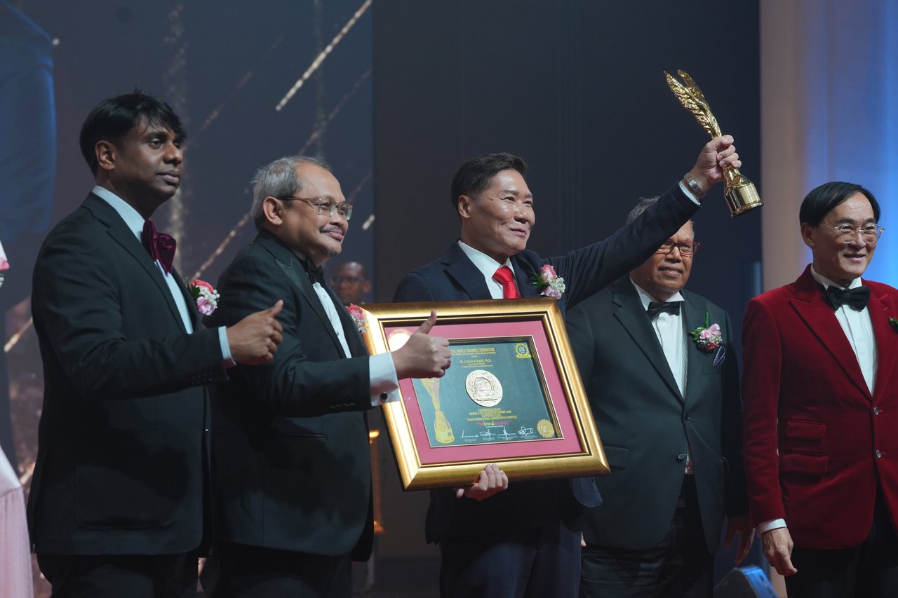 DR. J AARON K DAVID lifts the gold trophy and holds his certificate while Prof. Dr. Edward Roy Krishnan and a presenter beside him give a thumbs-up and Tun Md Raus Sharif stands behind him, seen from the side of the stage