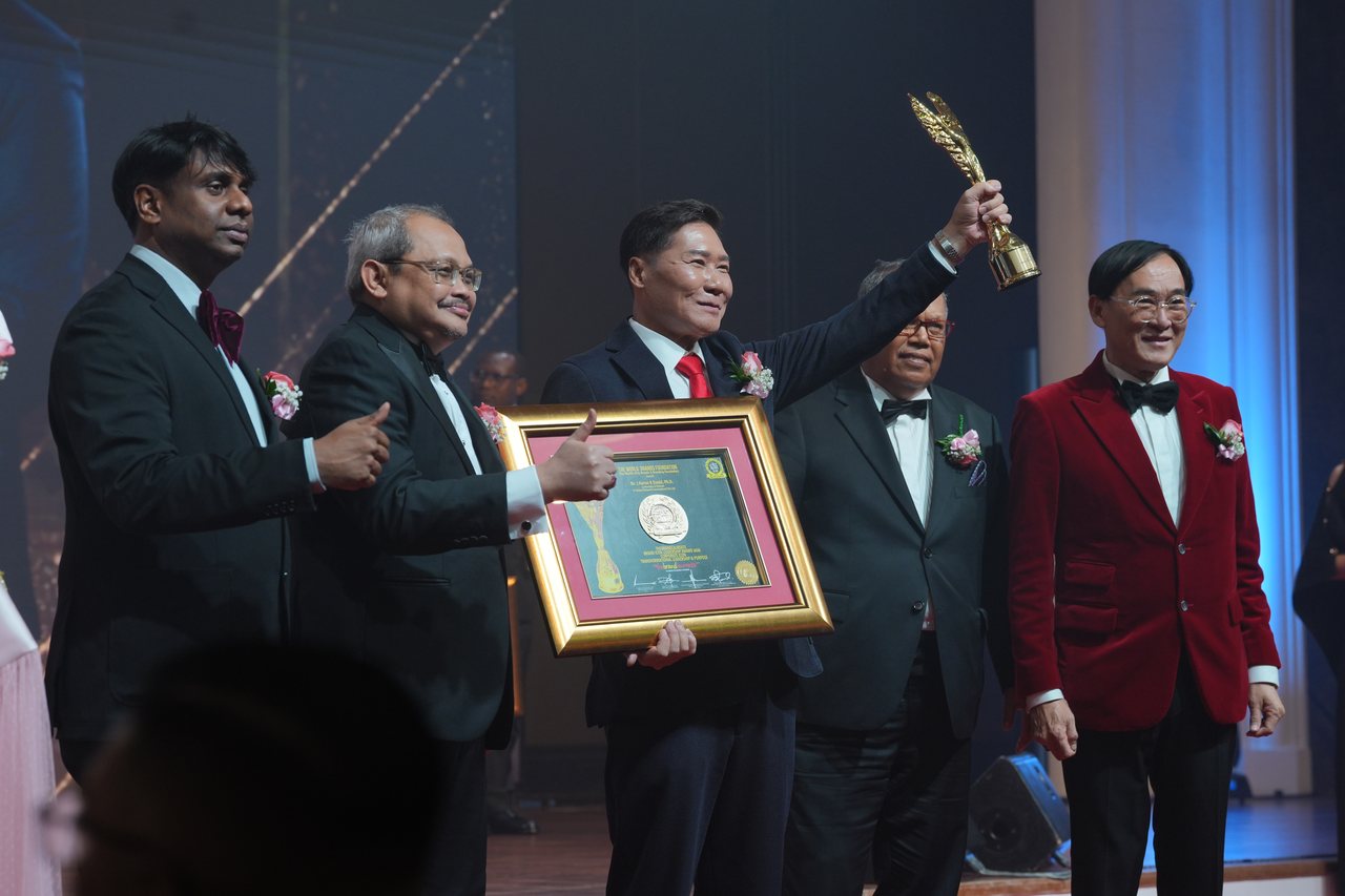 DR. J AARON K DAVID raises the gold trophy and holds his certificate while Prof. Dr. Edward Roy Krishnan and a presenter beside him give a thumbs-up, with Tun Md Raus Sharif behind him and Prof. Dr. KKJohan in a red velvet dinner jacket at right