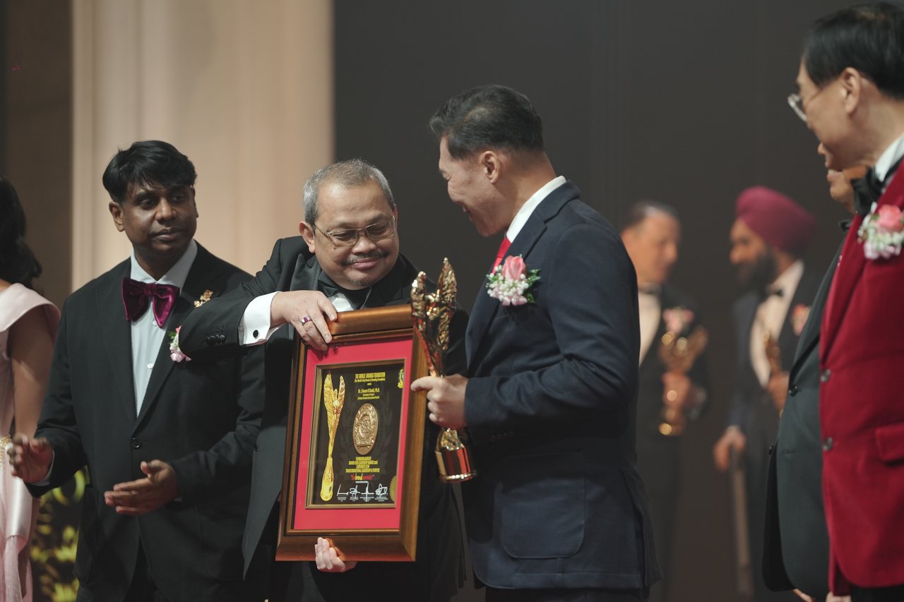 A presenter holds up the framed certificate while DR. J AARON K DAVID holds the gold trophy and Prof. Dr. Edward Roy Krishnan applauds at left, seen from the side of the stage