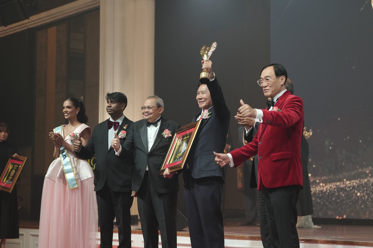 Seen from the side of the stage, left to right, Miss Universe Malaysia in a pale pink tulle gown and sash applauding, Prof. Dr. Edward Roy Krishnan and a presenter giving a thumbs-up, DR. J AARON K DAVID raising the gold trophy and holding his framed certificate, and Prof. Dr. KKJohan in a red velvet jacket giving a thumbs-up.