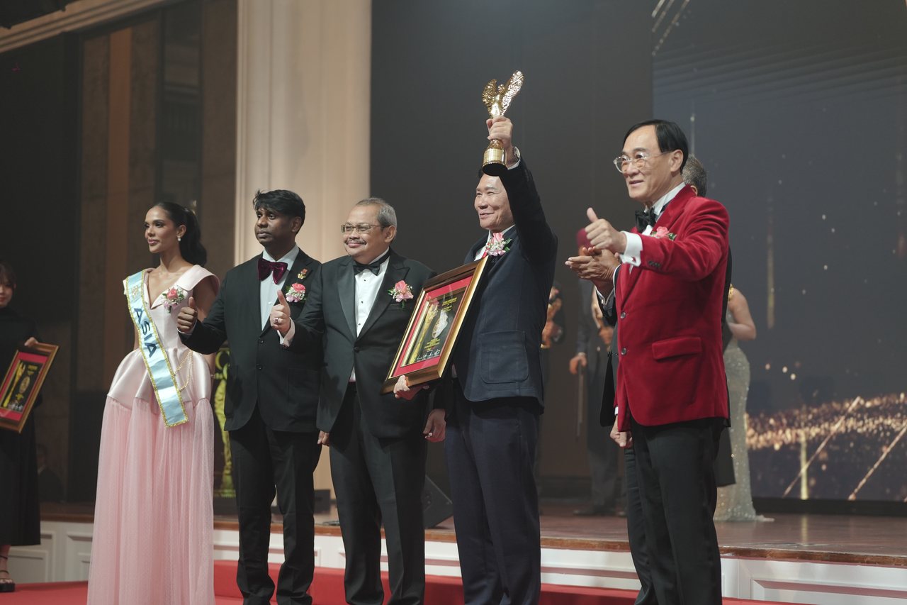 DR. J AARON K DAVID lifts the gold trophy and holds his framed certificate on stage while Prof. Dr. Edward Roy Krishnan, a presenter and Prof. Dr. KKJohan in a red velvet jacket give a thumbs-up beside him