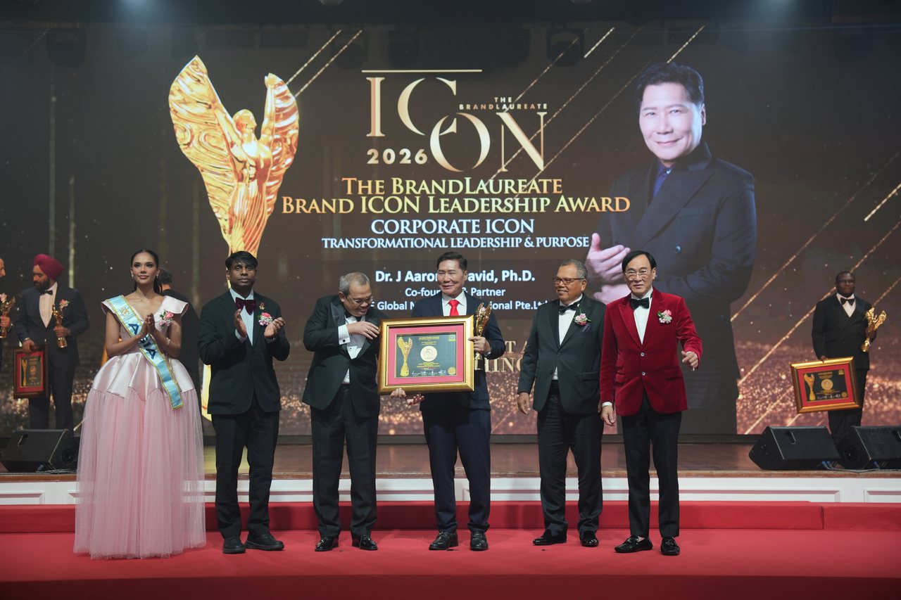 At centre stage DR. J AARON K DAVID holds the trophy and the framed certificate with a presenter, while Miss Universe Malaysia and Prof. Dr. Edward Roy Krishnan applaud at left and Tun Md Raus Sharif and a smiling Prof. Dr. KKJohan in a red velvet jacket stand at right, beneath the Corporate Icon screen with his name and portrait.
