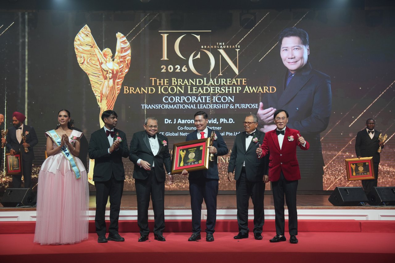 Left to right, Miss Universe Malaysia and Prof. Dr. Edward Roy Krishnan applauding, a presenter with his hands clasped, DR. J AARON K DAVID in a red tie holding the framed certificate and the gold trophy, Tun Md Raus Sharif standing beside him, and Prof. Dr. KKJohan in a red velvet jacket giving a thumbs-up, beneath the screen announcing Corporate Icon, Transformational Leadership and Purpose.