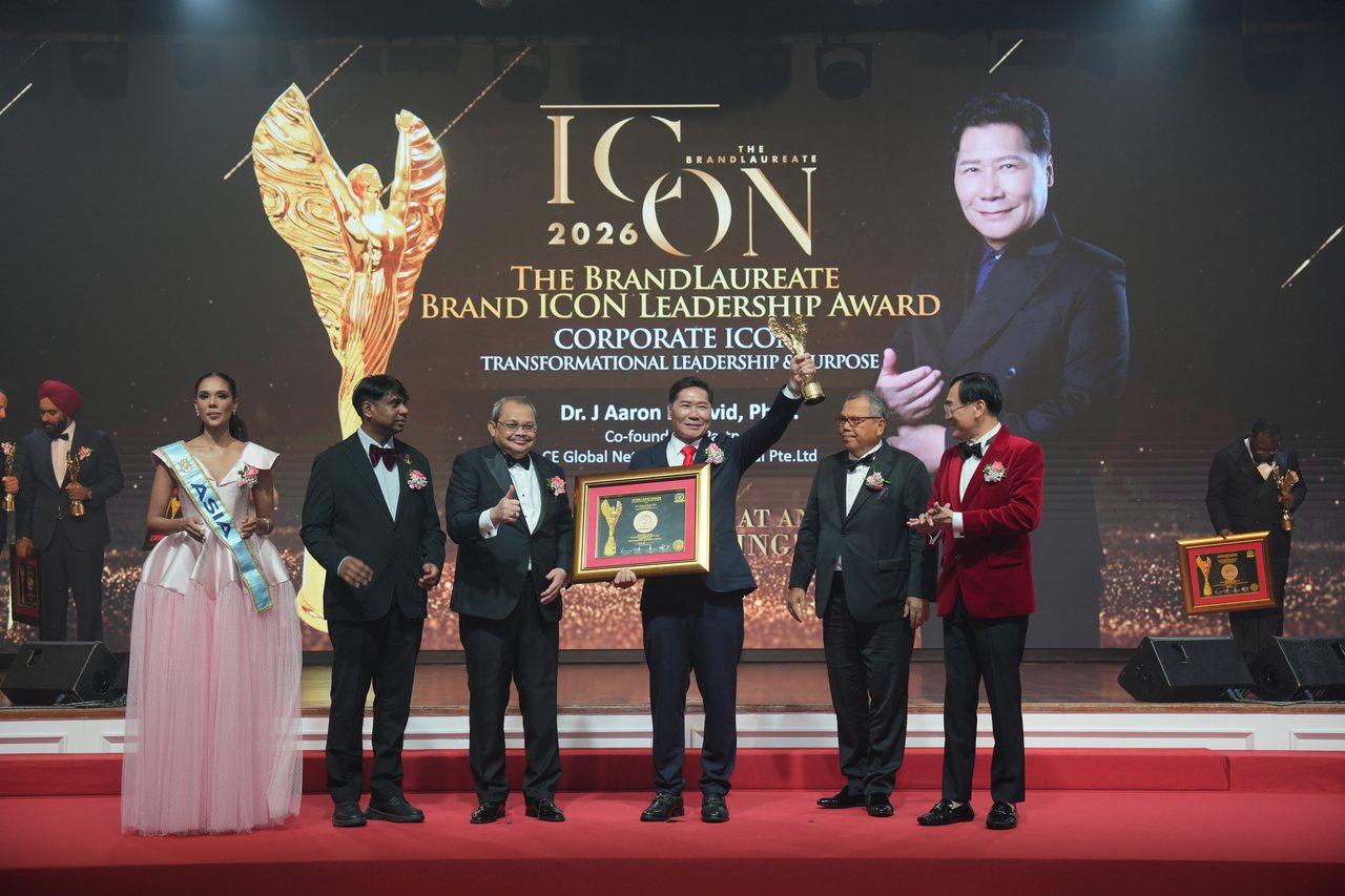 The full stage with DR. J AARON K DAVID at the centre raising his trophy, Prof. Dr. Edward Roy Krishnan looking toward him and a presenter giving a thumbs-up on the left, Tun Md Raus Sharif standing beside him on the right and Prof. Dr. KKJohan in a red velvet jacket, beneath the large screen with his portrait and award title