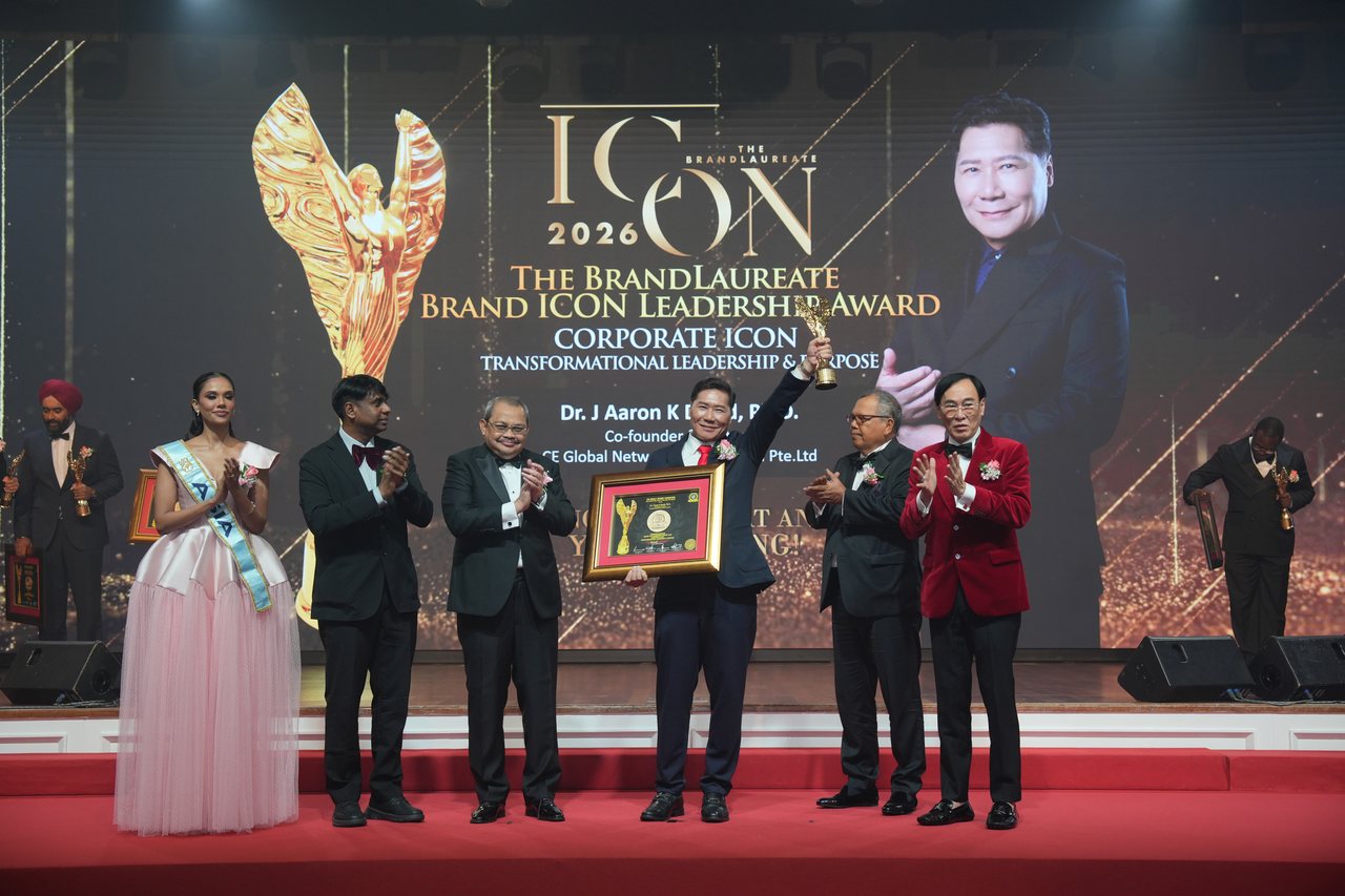 On the red-carpeted stage, left to right, Miss World Asia in a pale pink tulle gown with her sash, Prof. Dr. Edward Roy Krishnan and a presenter applauding, DR. J AARON K DAVID at the centre raising the gold winged trophy and holding his framed certificate, Tun Md Raus Sharif and Prof. Dr. KKJohan in his red velvet jacket applauding, beneath the giant screen with his portrait and the title Corporate Icon: Transformational Leadership & Purpose.