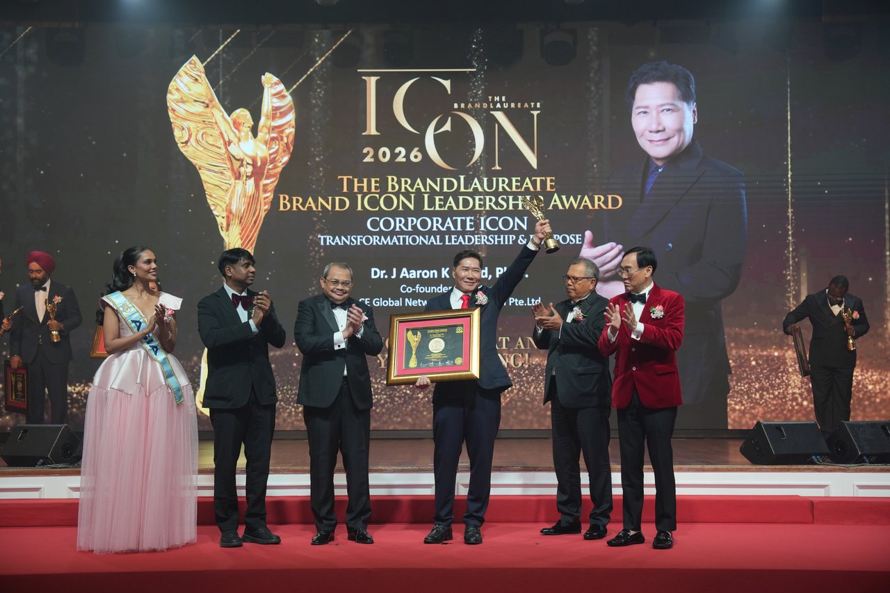 The full stage with DR. J AARON K DAVID at the centre raising his trophy and holding his certificate as Tun Md Raus Sharif, Prof. Dr. KKJohan, Prof. Dr. Edward Roy Krishnan, a presenter and Miss World Asia applaud, beneath the screen with his portrait and award title