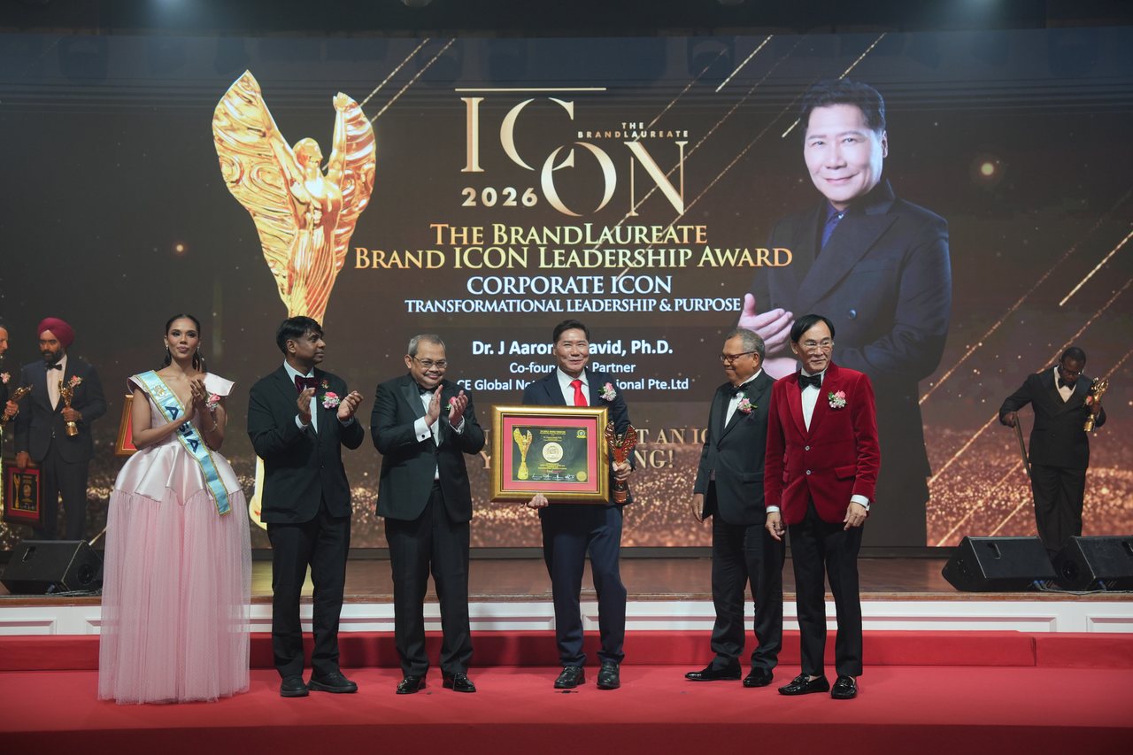DR. J AARON K DAVID faces the camera at centre stage with the framed certificate and gold winged trophy held before him, Miss World Asia, Prof. Dr. Edward Roy Krishnan and a presenter applauding on the left and Tun Md Raus Sharif and Prof. Dr. KKJohan in his red velvet jacket standing beside him on the right, beneath the screen reading The BrandLaureate Brand ICON Leadership Award, Corporate Icon.
