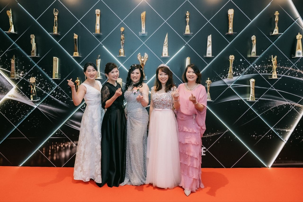 Five women in evening gowns stand on the orange carpet before a black wall of lit glass shelves holding gold trophies, left to right a guest in a white floral gown, a guest in a black gown with lace sleeves, DR. SHEKINAIH SIEW SIN YAP in her silver beaded gown with a pink corsage at the centre, a guest in a sparkling pink tulle gown with a tiara and a guest in a pink ruffled gown, all making finger-heart gestures.