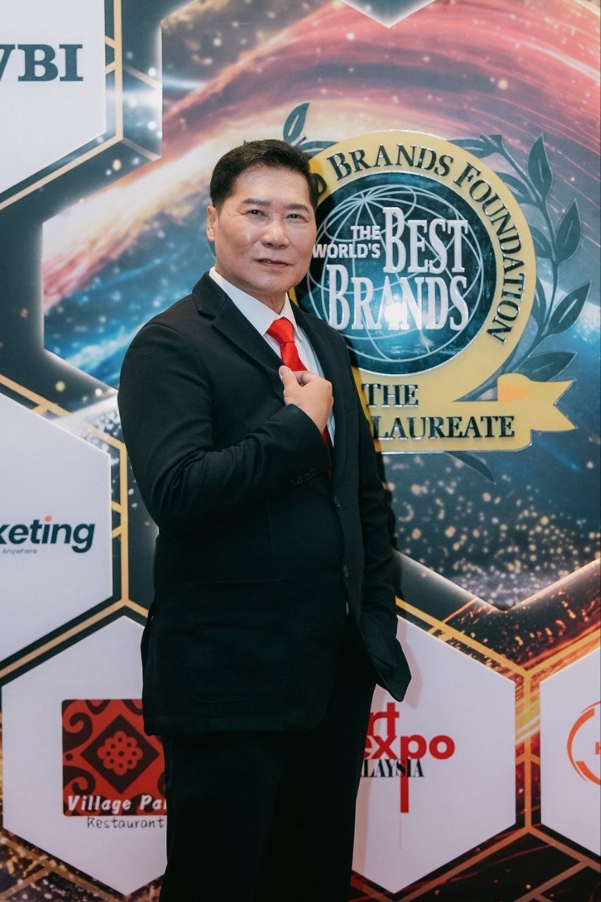DR. J AARON K DAVID, in a dark suit, white shirt and red tie, stands with one hand at his lapel before the gold World's Best Brands emblem of The World Brands Foundation and The BrandLaureate on the hexagon sponsor wall.