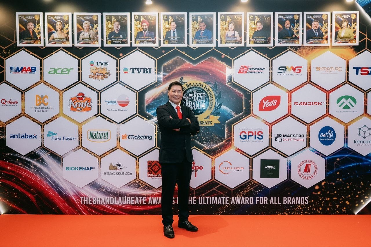 DR. J AARON K DAVID, arms folded, stands on the red carpet before the hexagon sponsor wall of The BrandLaureate, a row of framed magazine covers above