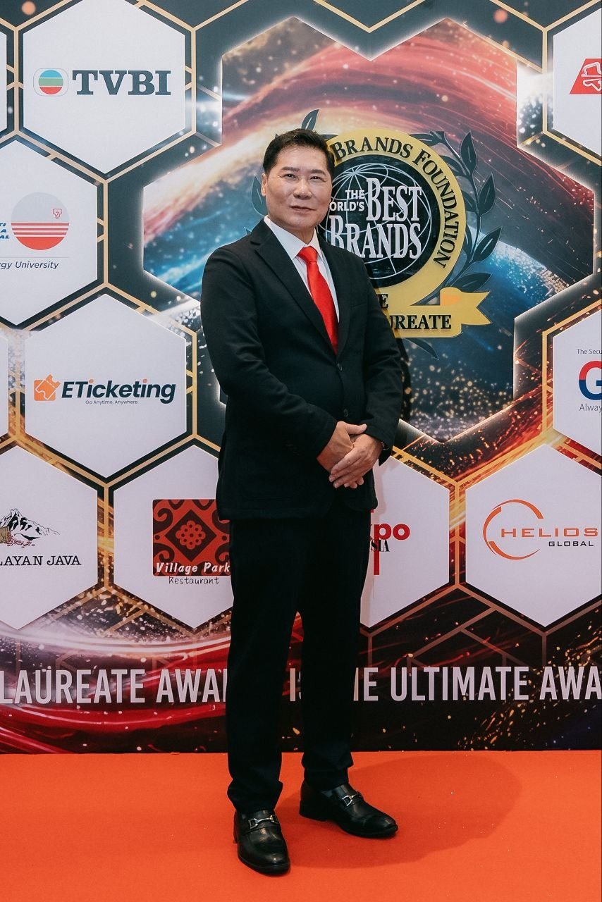 DR. J AARON K DAVID, in a navy suit and red tie, stands full-length on the red carpet before the hexagon sponsor wall of The World Brands Foundation and The BrandLaureate