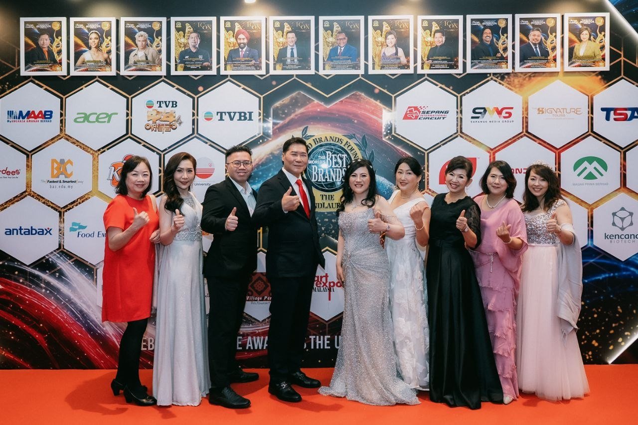 DR. J AARON K DAVID and DR. SHEKINAIH SIEW SIN YAP at the centre of a group of guests on the red carpet before the hexagon sponsor wall of The BrandLaureate, several giving a thumbs-up