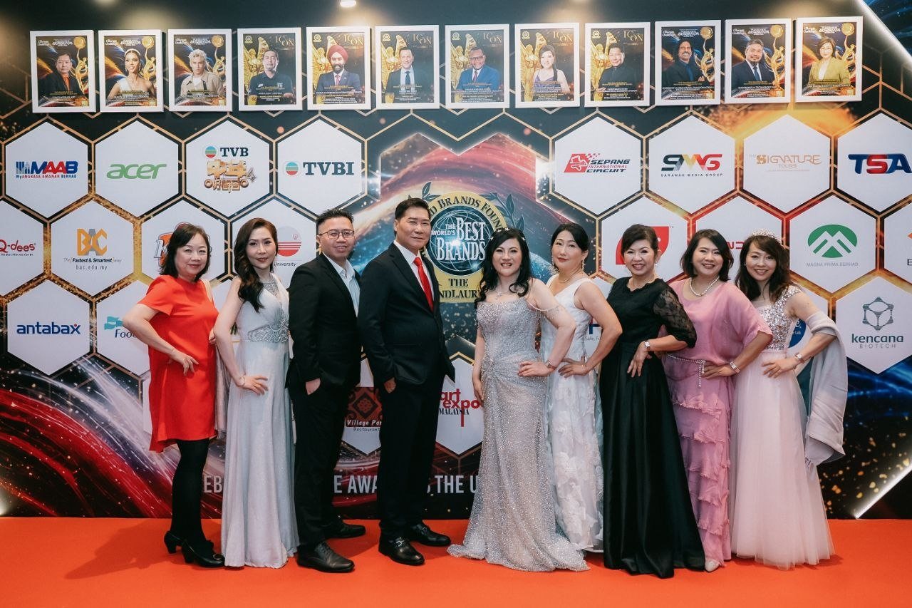Nine people in evening wear stand in a line on the orange carpet before the hexagon sponsor wall of The BrandLaureate, DR. J AARON K DAVID in his red tie and DR. SHEKINAIH SIEW SIN YAP in her silver beaded gown at the centre, guests in red, pale silver, black, white, black lace, pink and pale pink on either side, with framed ICON magazine covers of the recipients along the top.