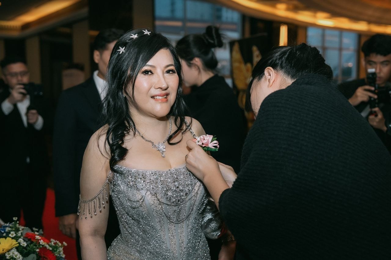 DR. SHEKINAIH SIEW SIN YAP smiles at the camera as a pink rose corsage is pinned to the strap of her silver beaded gown, photographers behind her