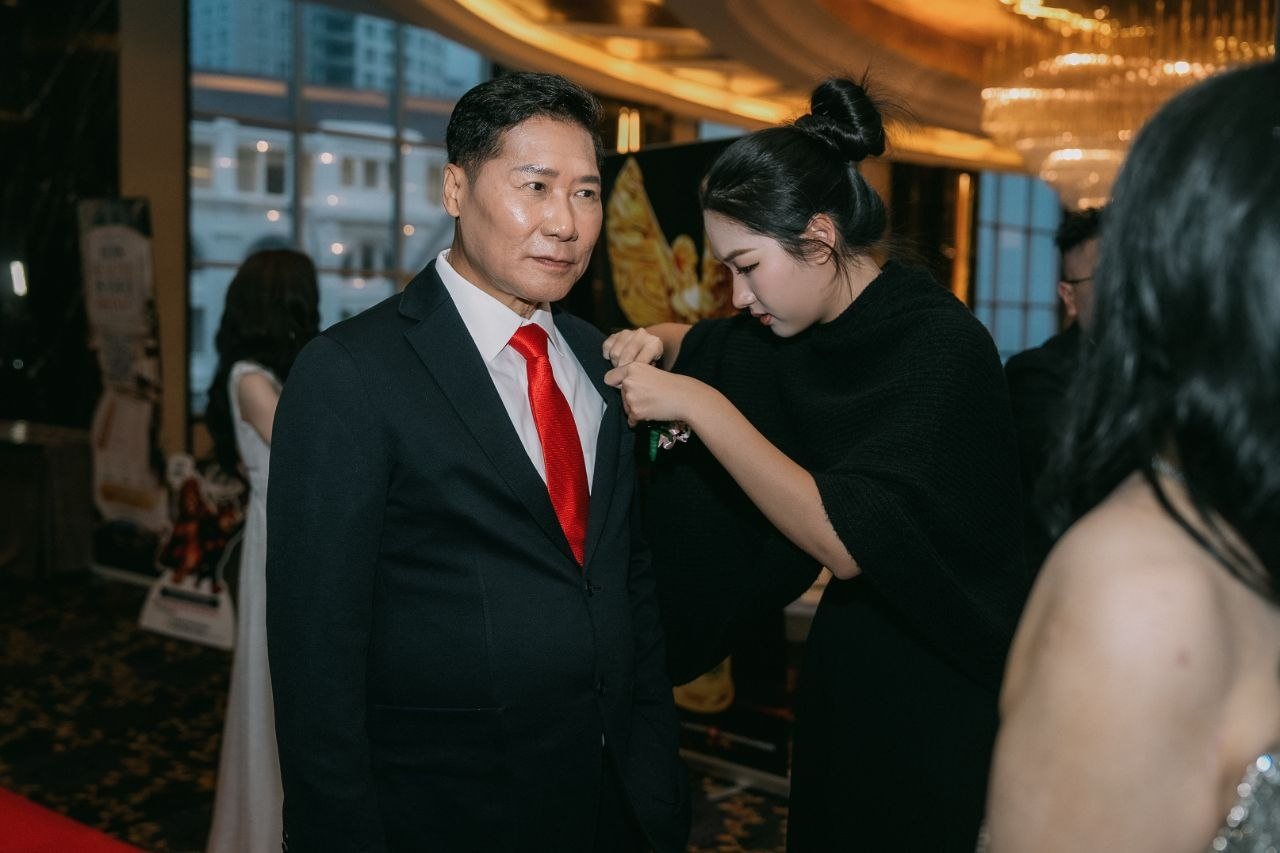 DR. J AARON K DAVID, in a dark suit and red tie, stands calmly as a guest in a black knit wrap pins a corsage to his lapel, the hotel windows and a golden trophy banner behind them.