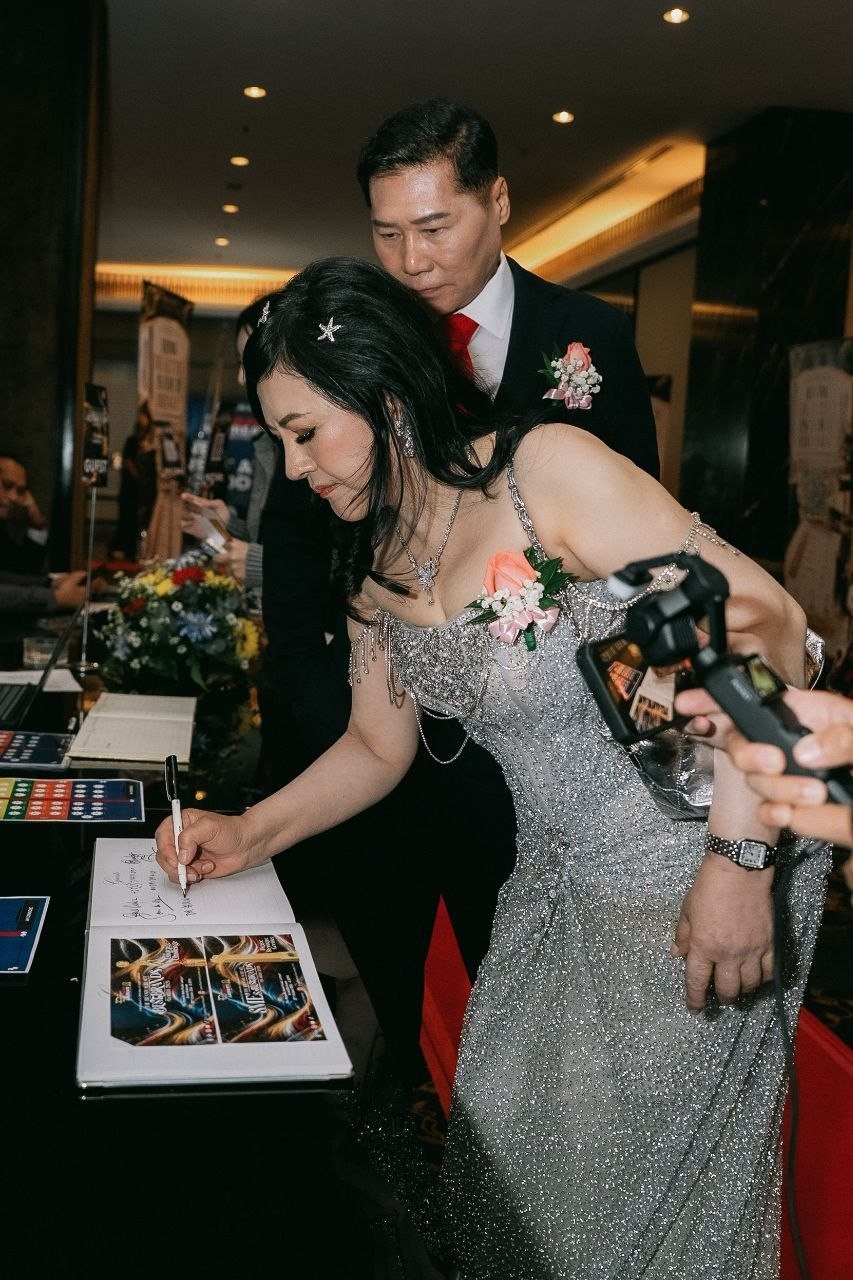 DR. SHEKINAIH SIEW SIN YAP, in her silver beaded gown with a pink rose corsage, bends over the guestbook at the reception table with a pen in hand, DR. J AARON K DAVID standing close behind her and a guest filming on a phone gimbal at the right.
