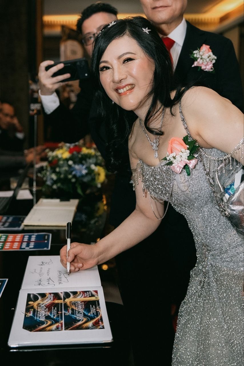DR. SHEKINAIH SIEW SIN YAP leans over the guestbook at the reception table with a pen in hand, looking up and laughing, as DR. J AARON K DAVID stands behind her and a guest photographs on a phone