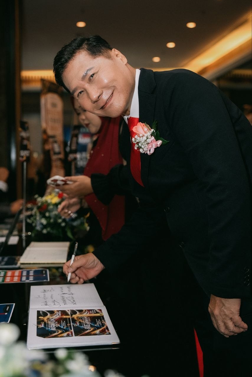 DR. J AARON K DAVID leans over the guestbook at the reception table with a pen in hand, smiling at the camera