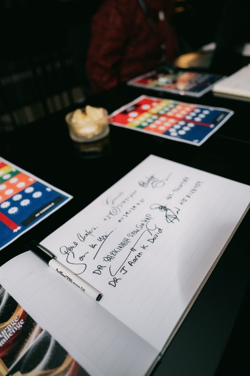 A close view of the open guestbook on the dark reception table, with the handwritten entries and signatures of DR. SHEKINAIH SIEW SIN YAP and DR. J AARON K DAVID, a marker pen resting on the page and colourful table cards beyond.