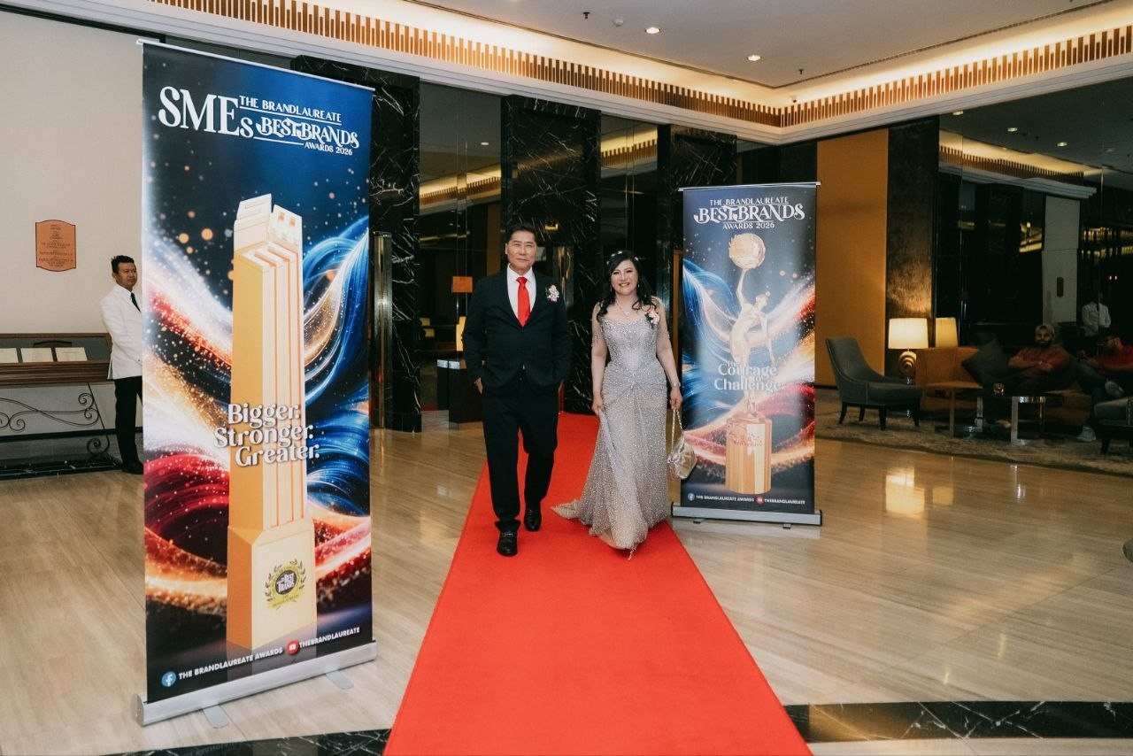 DR. J AARON K DAVID and DR. SHEKINAIH SIEW SIN YAP walk side by side along the red carpet across the marble lobby, between the two award banners