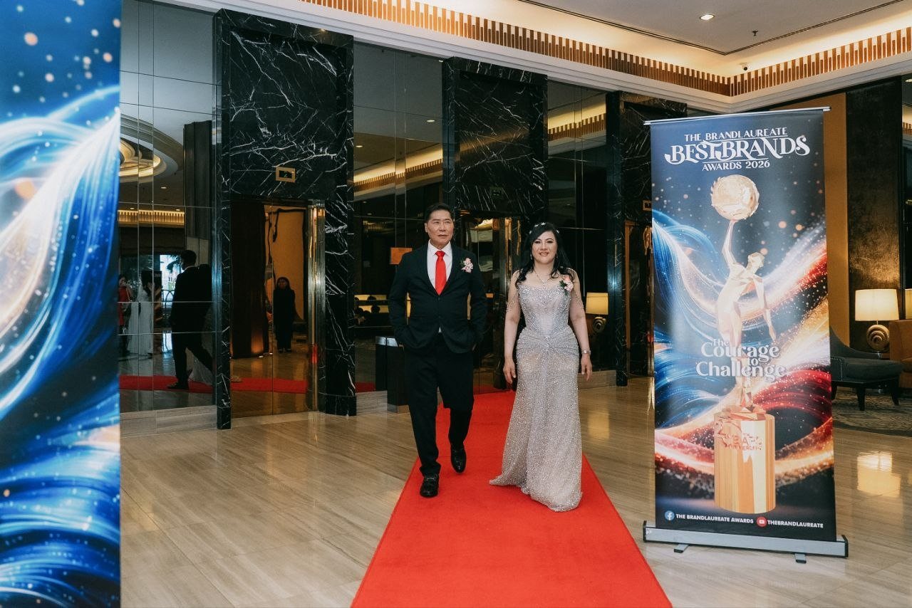 DR. J AARON K DAVID and DR. SHEKINAIH SIEW SIN YAP walk side by side down the red carpet from the black marble lift lobby, the banner of The BrandLaureate BestBrands Awards 2026 standing at the right.