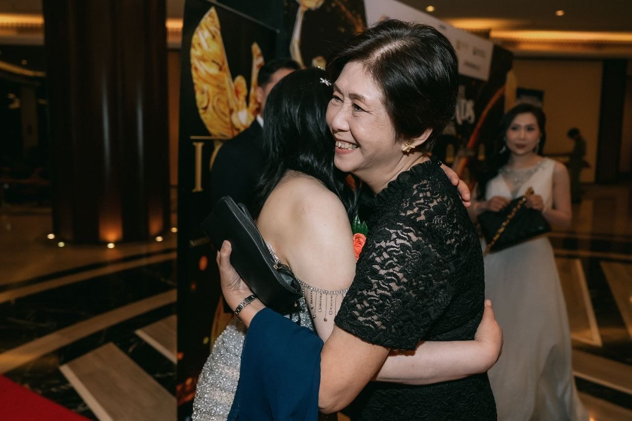 A key member of The BrandLaureate team, in a black lace dress, smiles as she embraces DR. SHEKINAIH SIEW SIN YAP, seen from behind in her beaded silver gown, while Florence, in an ivory sleeveless V-neck gown with a dark clutch bag, stands smiling in the background.