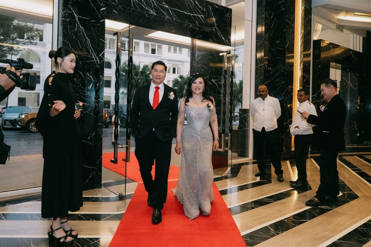 DR. J AARON K DAVID and DR. SHEKINAIH SIEW SIN YAP walk the red carpet just inside the hotel’s glass doors, a camera operator and guests on either side