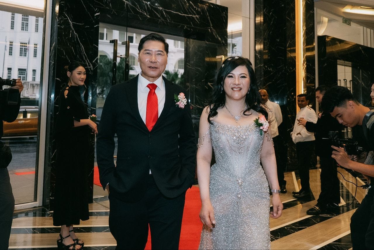 DR. SHEKINAIH SIEW SIN YAP and DR. J AARON K DAVID walk in on a red carpet through tall glass doors in black marble frames, white colonial-style buildings across the street behind them