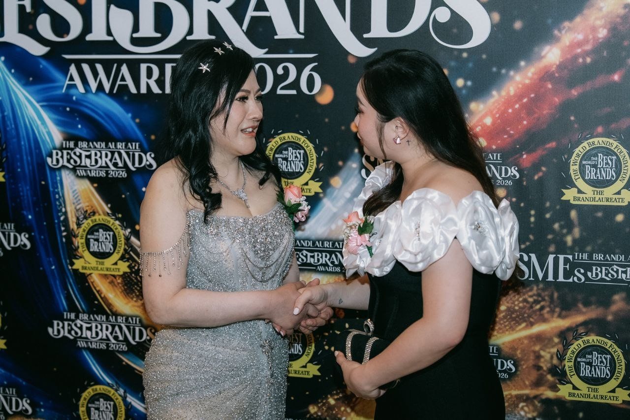 DR. SHEKINAIH SIEW SIN YAP smiles and shakes hands with Hilary Ng, in a black gown with white rose sleeves, before The BrandLaureate BestBrands Awards 2026 backdrop