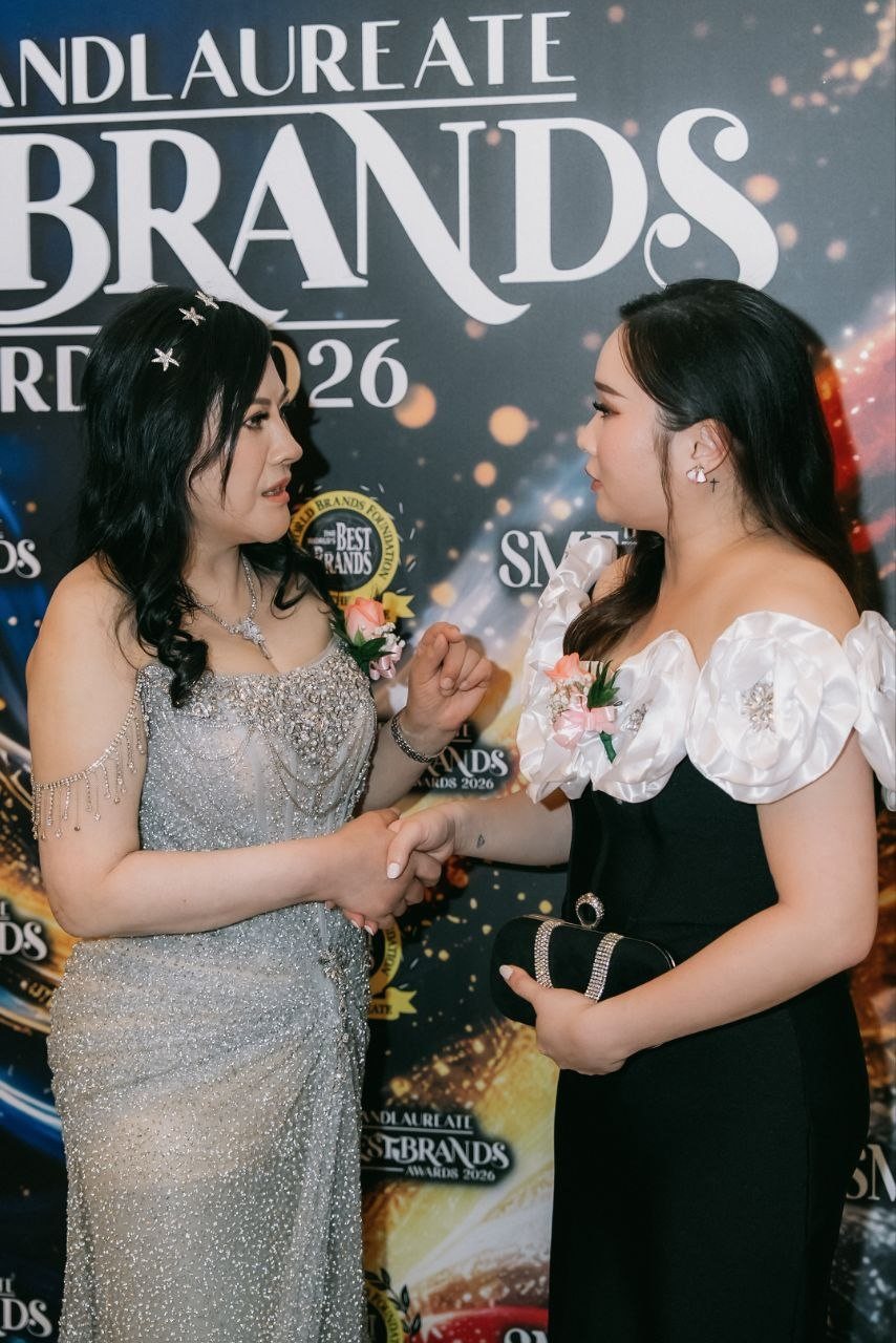 DR. SHEKINAIH SIEW SIN YAP, in her silver beaded gown, and Hilary Ng, in a black gown with white rose sleeves and holding a black clutch, clasp hands and talk before The BrandLaureate BestBrands Awards 2026 backdrop.