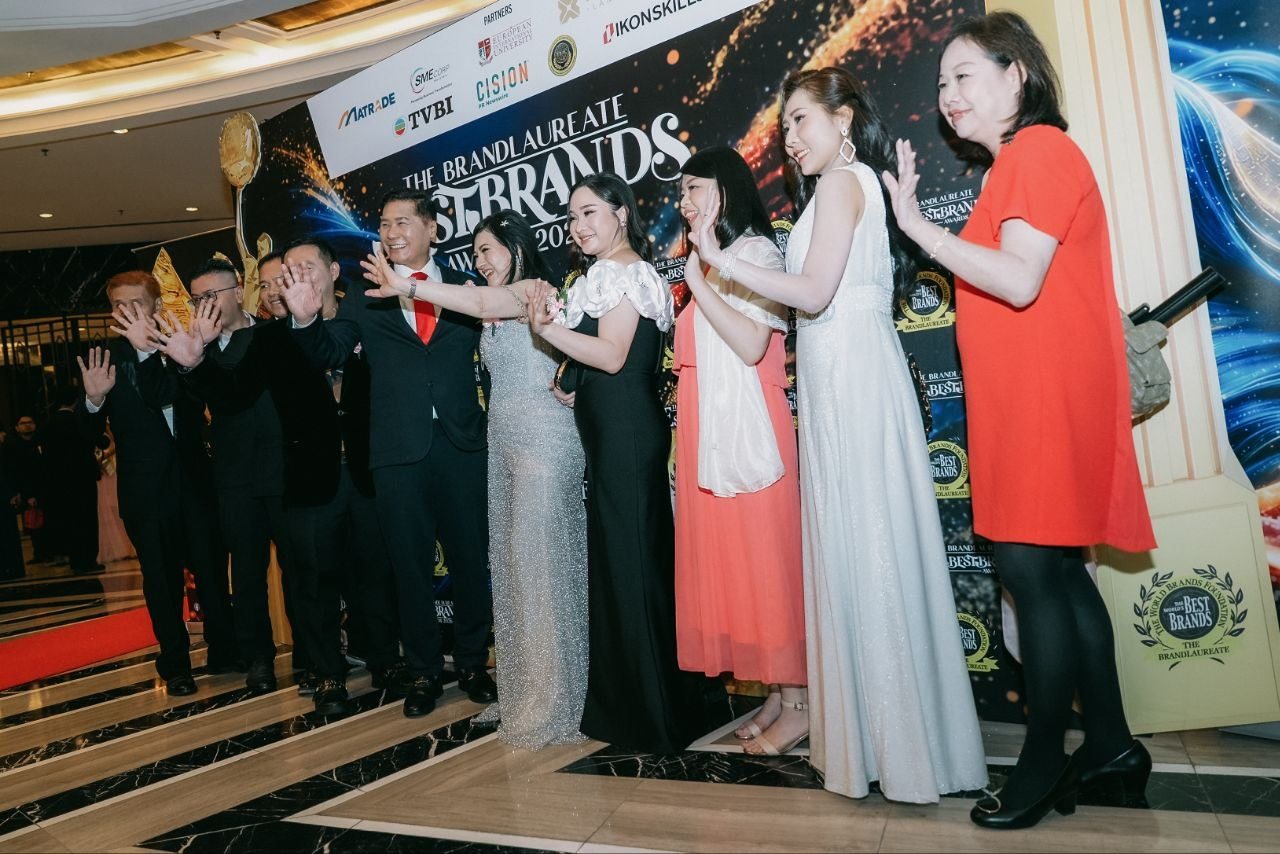 Seen from a low side angle, DR. J AARON K DAVID, DR. SHEKINAIH SIEW SIN YAP, Hilary Ng, a guest in coral, Florence in her ivory sleeveless gown and a guest in red wave toward the photographers alongside the team of CE Global Network International and Crowned Eagles Global at the BestBrands backdrop.