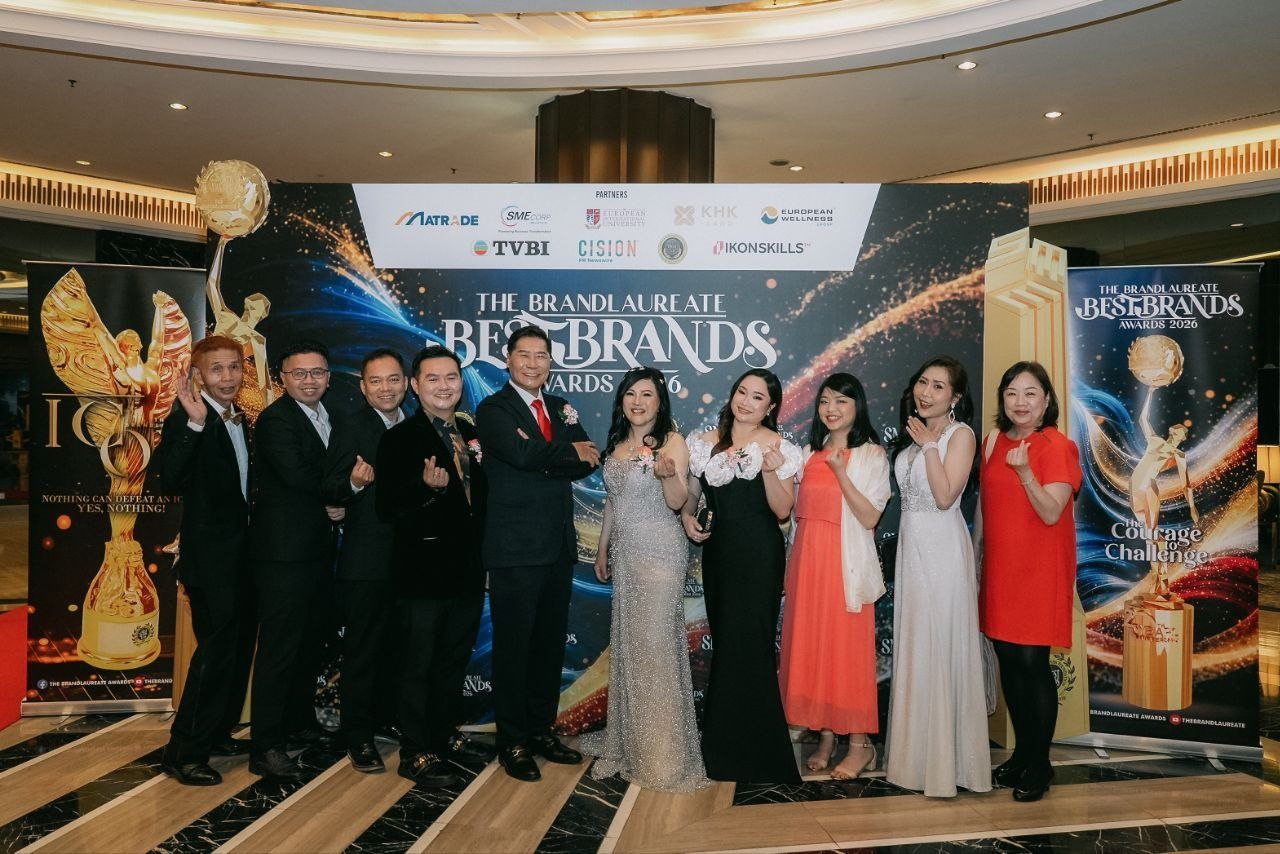 In a wide shot of the domed lobby, DR. J AARON K DAVID and DR. SHEKINAIH SIEW SIN YAP stand at the centre of a row with Hilary Ng, Florence in an ivory sleeveless gown and the team of CE Global Network International and Crowned Eagles Global, several making finger-heart gestures before The BrandLaureate BestBrands Awards 2026 backdrop.