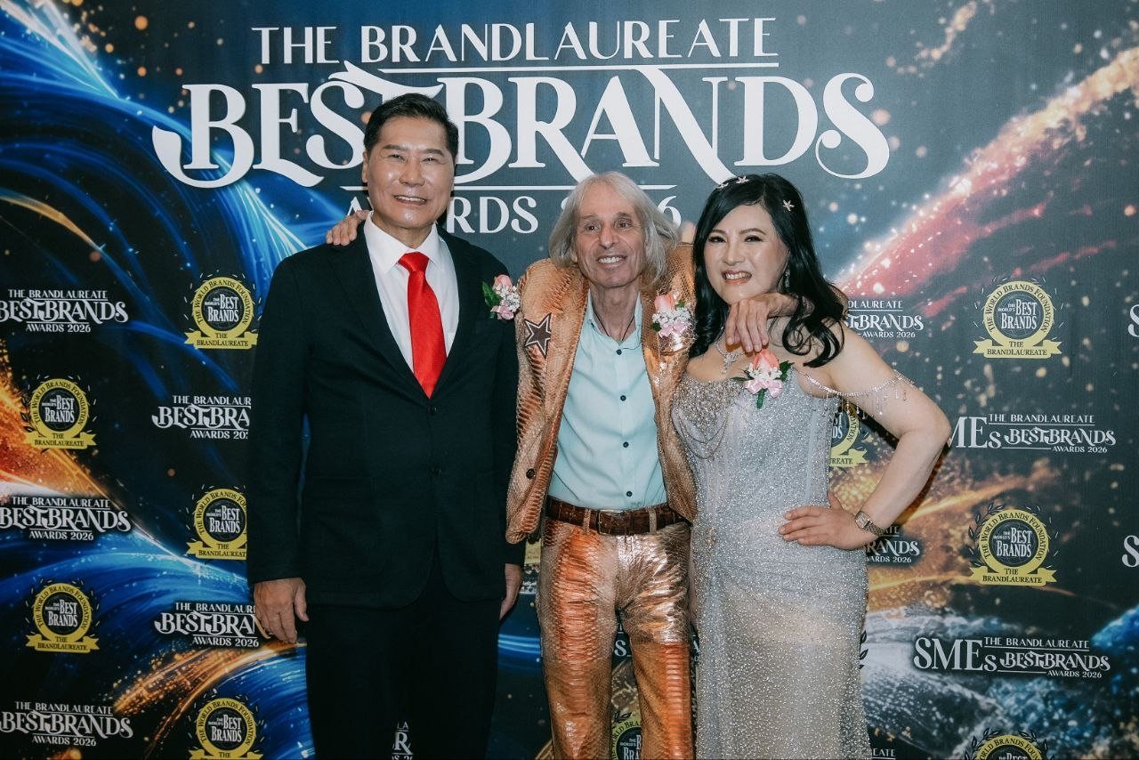 DR. J AARON K DAVID and DR. SHEKINAIH SIEW SIN YAP stand either side of Alain Robert, in a gold jacket and gold trousers, before The BrandLaureate BestBrands Awards 2026 backdrop