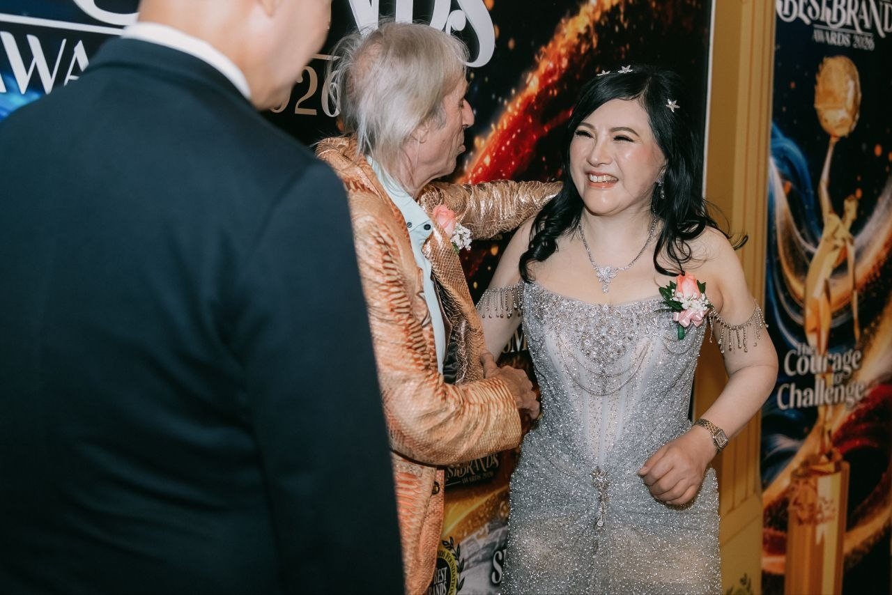 DR. SHEKINAIH SIEW SIN YAP laughs as Alain Robert, in his gold snakeskin jacket, leans in to greet her, a gentleman in a dark suit in the foreground with his back to the camera.
