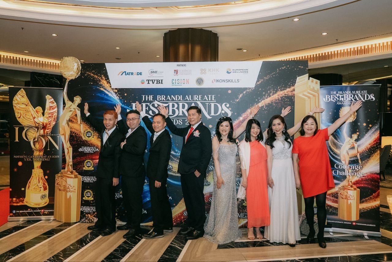 Left to right, three men of the team in black suits, DR. J AARON K DAVID in a navy suit and red tie, DR. SHEKINAIH SIEW SIN YAP in a silver beaded gown, a team member in a coral dress with a white shawl, Florence in an ivory beaded gown and a team member in a red dress raise their arms before The BrandLaureate BestBrands Awards 2026 backdrop, with a tall gold trophy replica on the left and a gold column on the right.